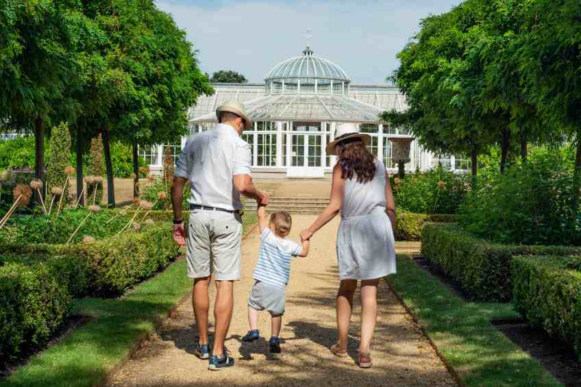 Paar mit Kind in der Mitte geht durch den Park auf ein Gewächshaus zu. Beau Pankow bietet familienfreundliche Eigentumswohnungen im grünen Berlin-Pankow.